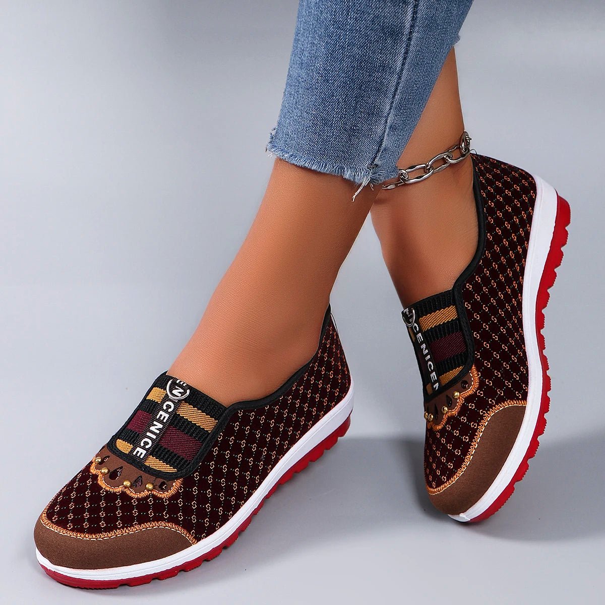 Brown patterned slip-on shoes with red soles worn with blue jeans on a gray background