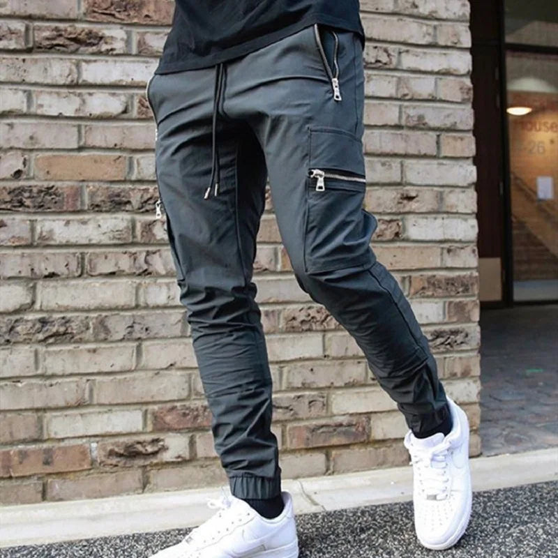 Person wearing dark gray jogger pants with zippers and white sneakers in front of a brick wall.