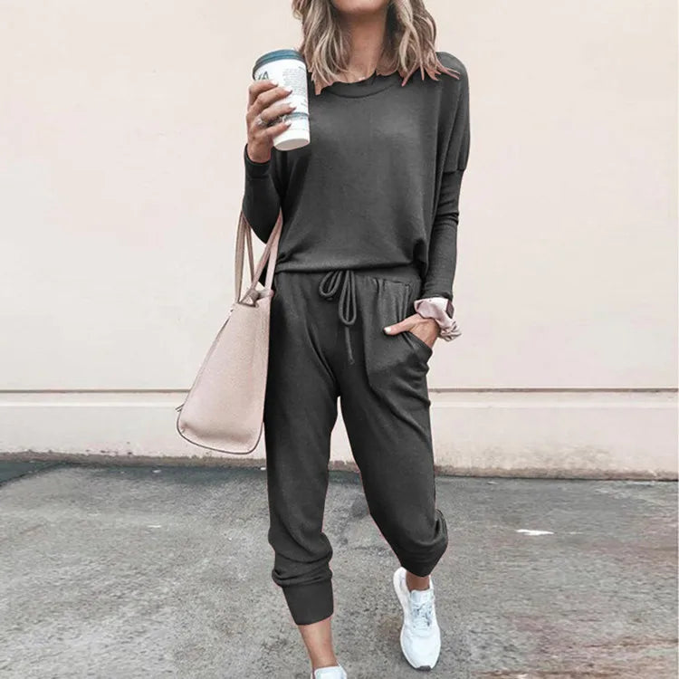 Person wearing a dark gray tracksuit with a beige handbag and white sneakers, standing against a light pink wall.