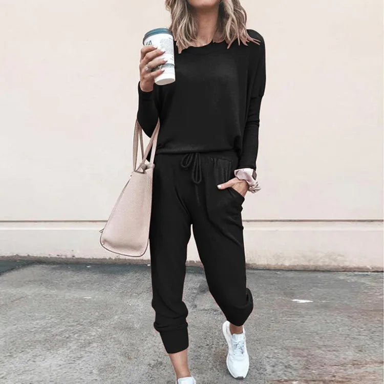 Person wearing a black outfit with a beige handbag and white sneakers, standing against a light pink wall.