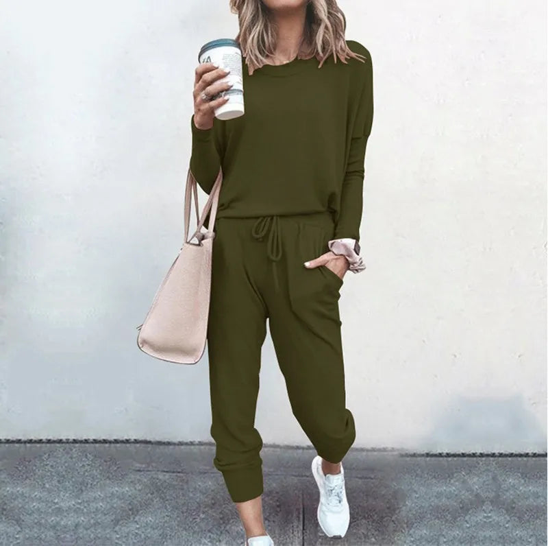 Person wearing an olive green outfit with a beige bag and white sneakers, standing against a light gray background.